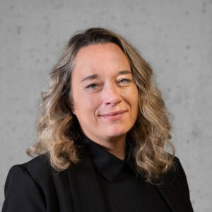 Eine Person mit schulterlangem, welligem Haar, die einen schwarzen Blazer und ein schwarzes Hemd trägt, lächelt leicht, während sie vor einem schlichten grauen Hintergrund steht.