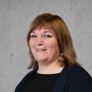 Eine Person mit schulterlangem hellbraunem Haar, einem schwarzen Oberteil und einer dunklen Jacke lächelt vor einem grauen Hintergrund. Sie hat große runde Ohrringe und blaue Augen.