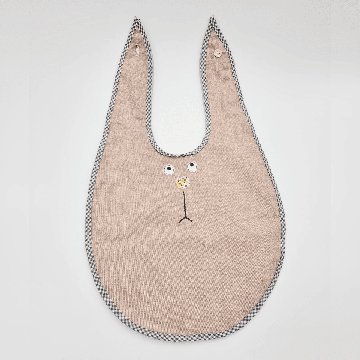 Das Esslätzchen ist ein beiges Lätzchen in Hasenform mit kariertem Rand und einem gestickten Gesicht auf einfarbigem Hintergrund.