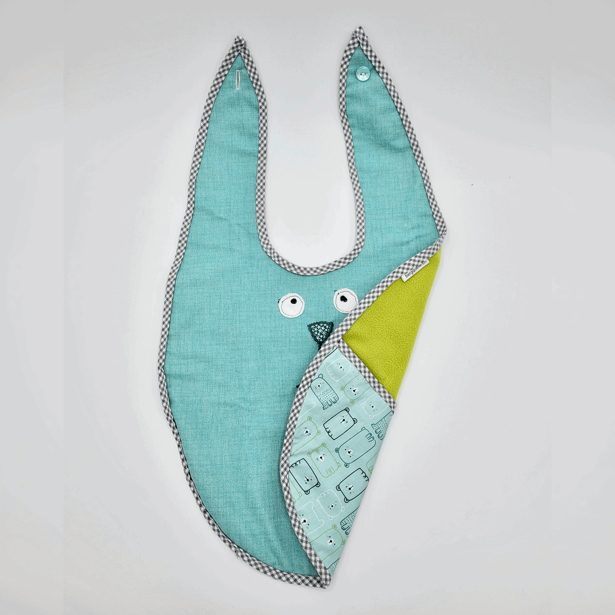 Das Esslätzchen ist ein Lätzchen in Hasenform mit einer türkisfarbenen Vorderseite, gemustertem Rand und einer gelben Wendeseite mit Muster.