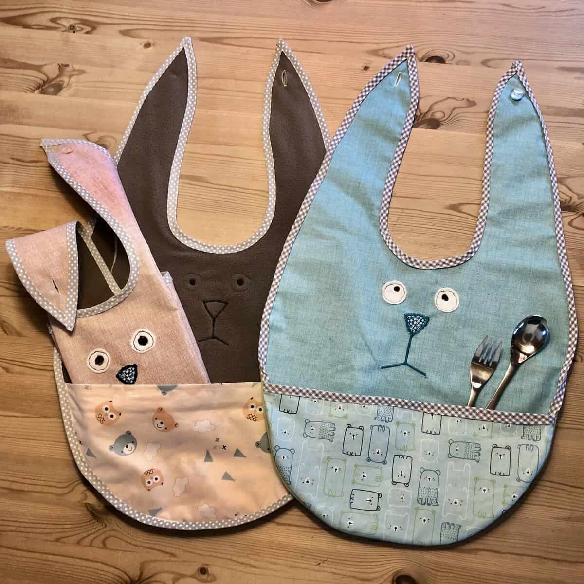 Auf einem Holztisch liegen drei Esslätzchen-Lätzchen mit Tiermotiven sowie Gabel und Löffel.
