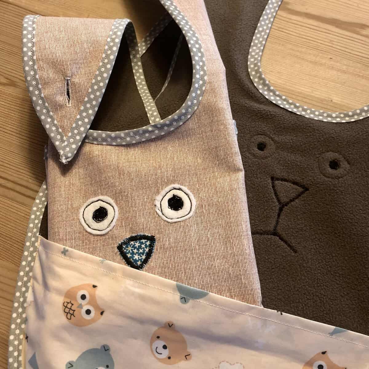 Zwei Esslätzchen-Stofflätzchen mit Tiergesichtern: eines braun mit Ohren und ein beige mit Eulenaugen und -nase, auf einer Holzoberfläche platziert.