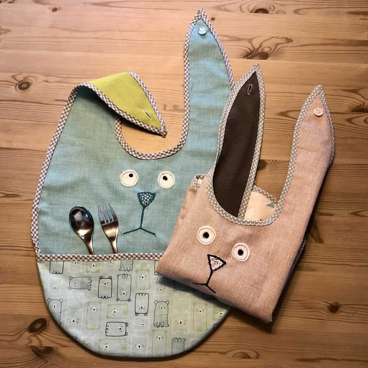 Auf einem Holztisch liegen zwei Esslätzchen-Lätzchen in Häschenform mit Bestecktasche.
