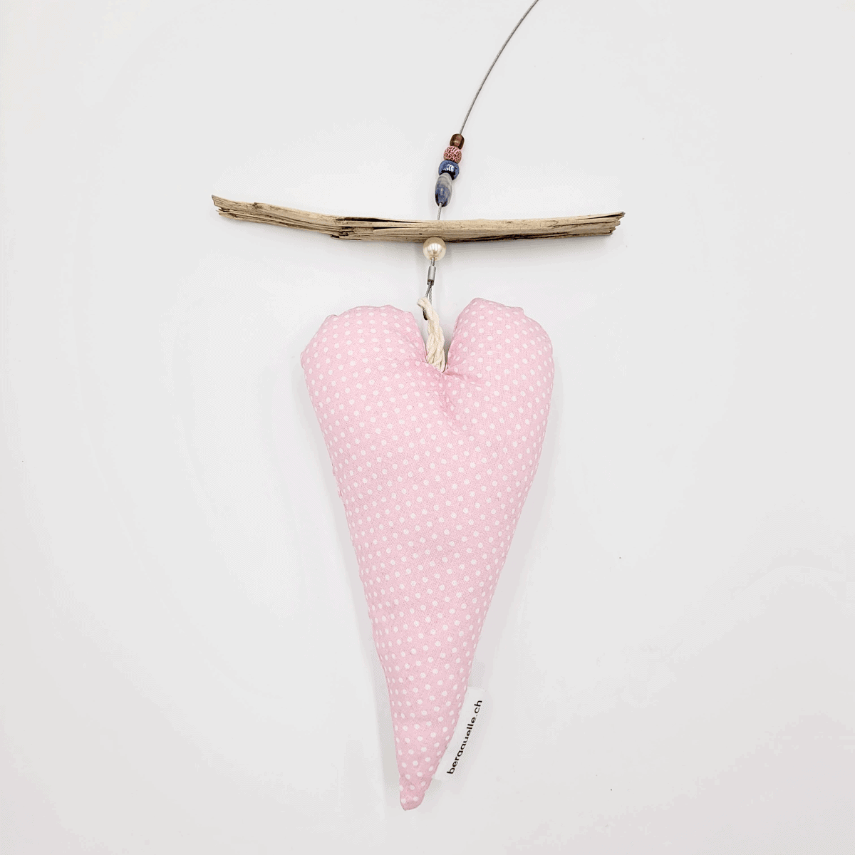 Eine „Fotoleine mit Herz“ zeigt ein herzförmiges Kissen mit rosa Punkten, das an Treibholz hängt, vor einem weißen Hintergrund.