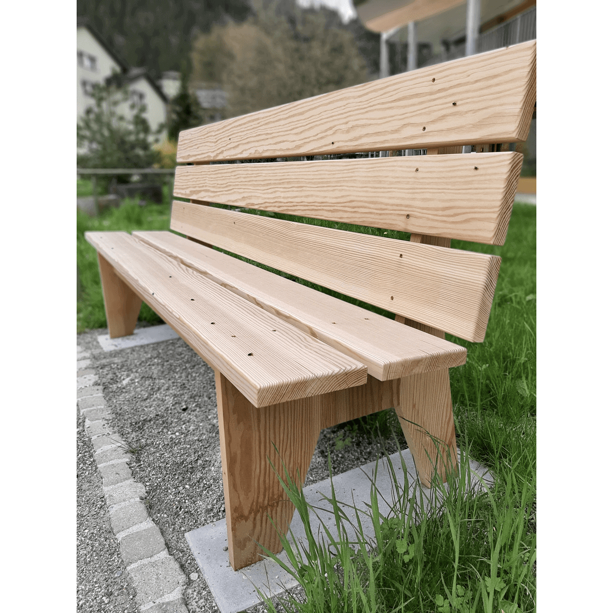 Holzbank auf einer Rasenfläche neben einem Steinweg, mit Gebäuden und Bäumen im Hintergrund.