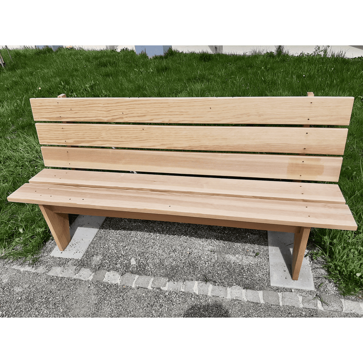 Die Holzbank steht auf einem Betonweg, im Hintergrund ist grünes Gras zu sehen.