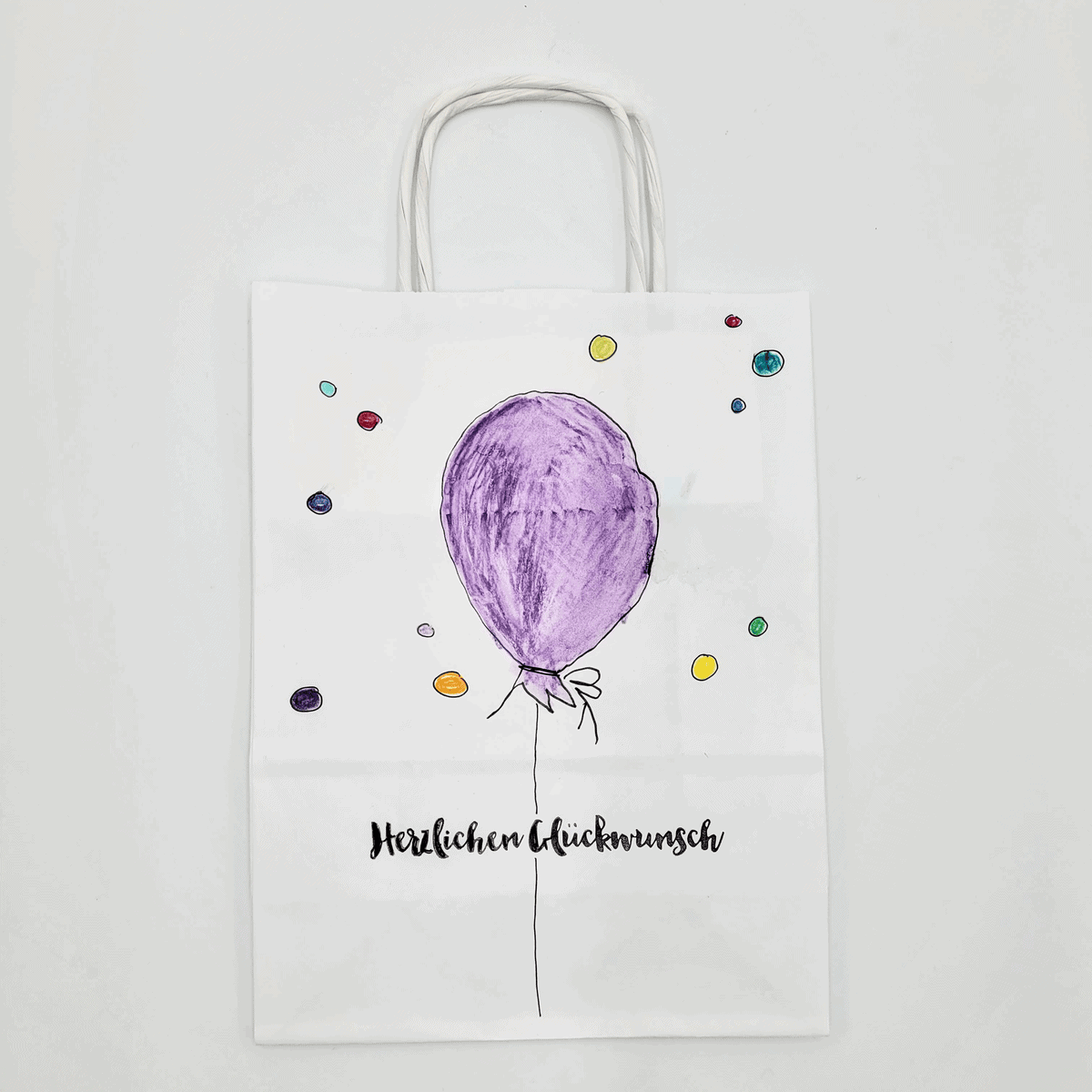 Papiertasche bemalt mit einem weißen Geschenktüten-Design mit einem lila Luftballon, bunten Punkten und dem Text „Herzlichen Glückwunsch“.
