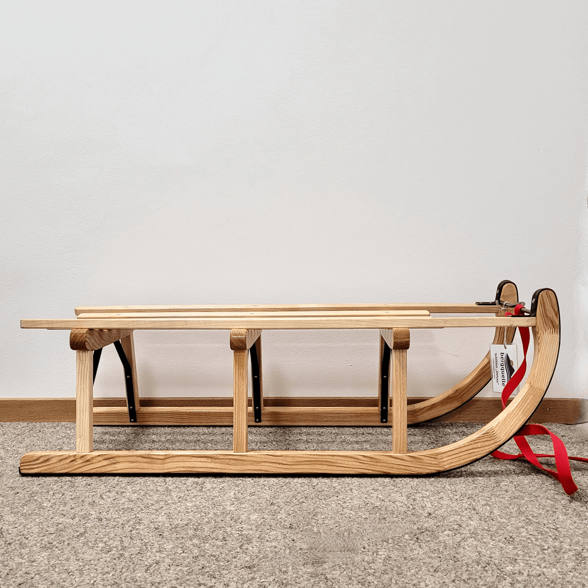 Der Schlitten mit rotem Riemen lehnt an einer weißen Wand auf einem Teppichboden.