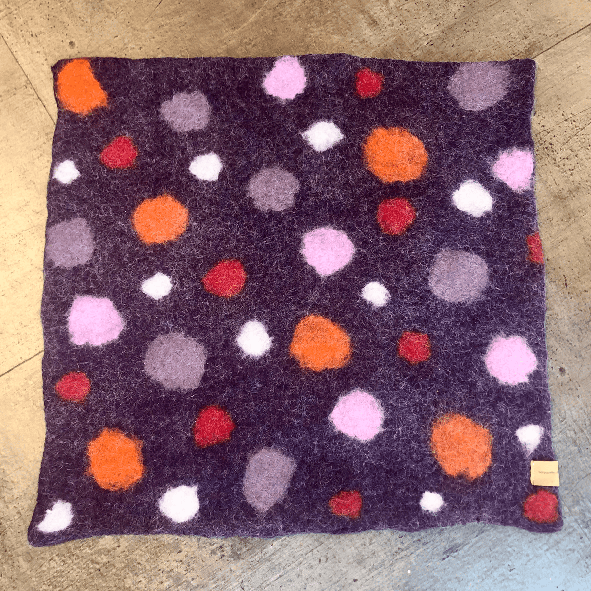 Ein Sitzkissen mit einem lila Stoffquadrat, verziert mit lebendigen kreisförmigen Mustern in Orange, Rosa, Weiß und Rot, auf einer Holzoberfläche platziert.