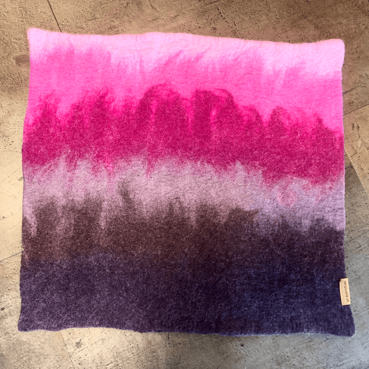 Ein quadratisches Sitzkissen aus Wollfilz weist einen Farbverlauf von Rosa nach Lila auf und ruht auf einem strukturierten Boden.