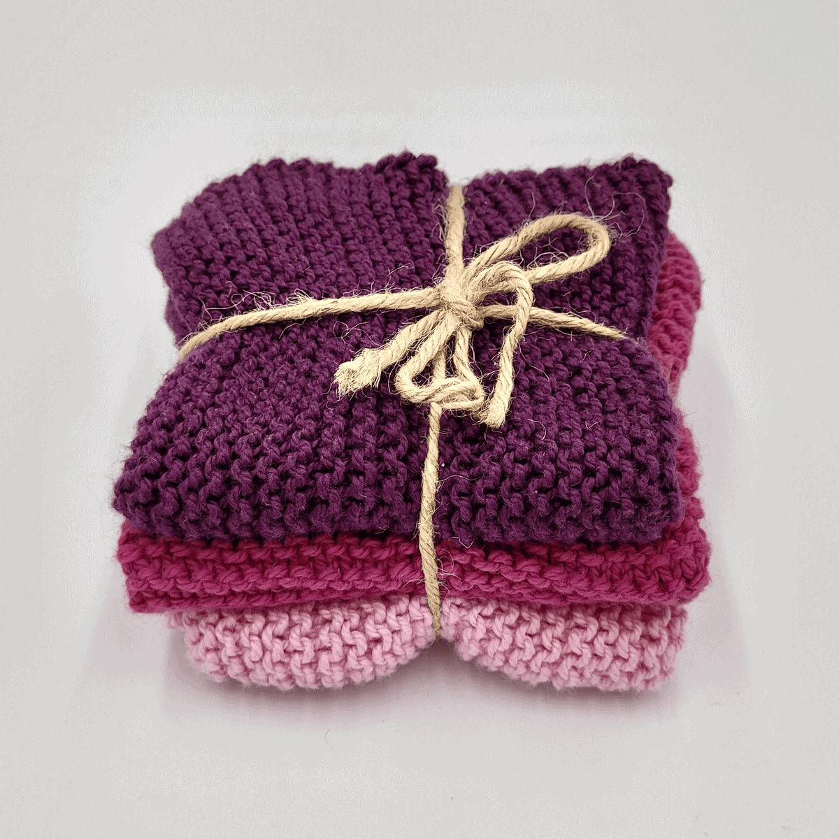 Auf weißem Untergrund liegt ein Stapel gestrickter Spüllappen in Lila und Rosa, zusammengebunden mit einer beigen Kordel.