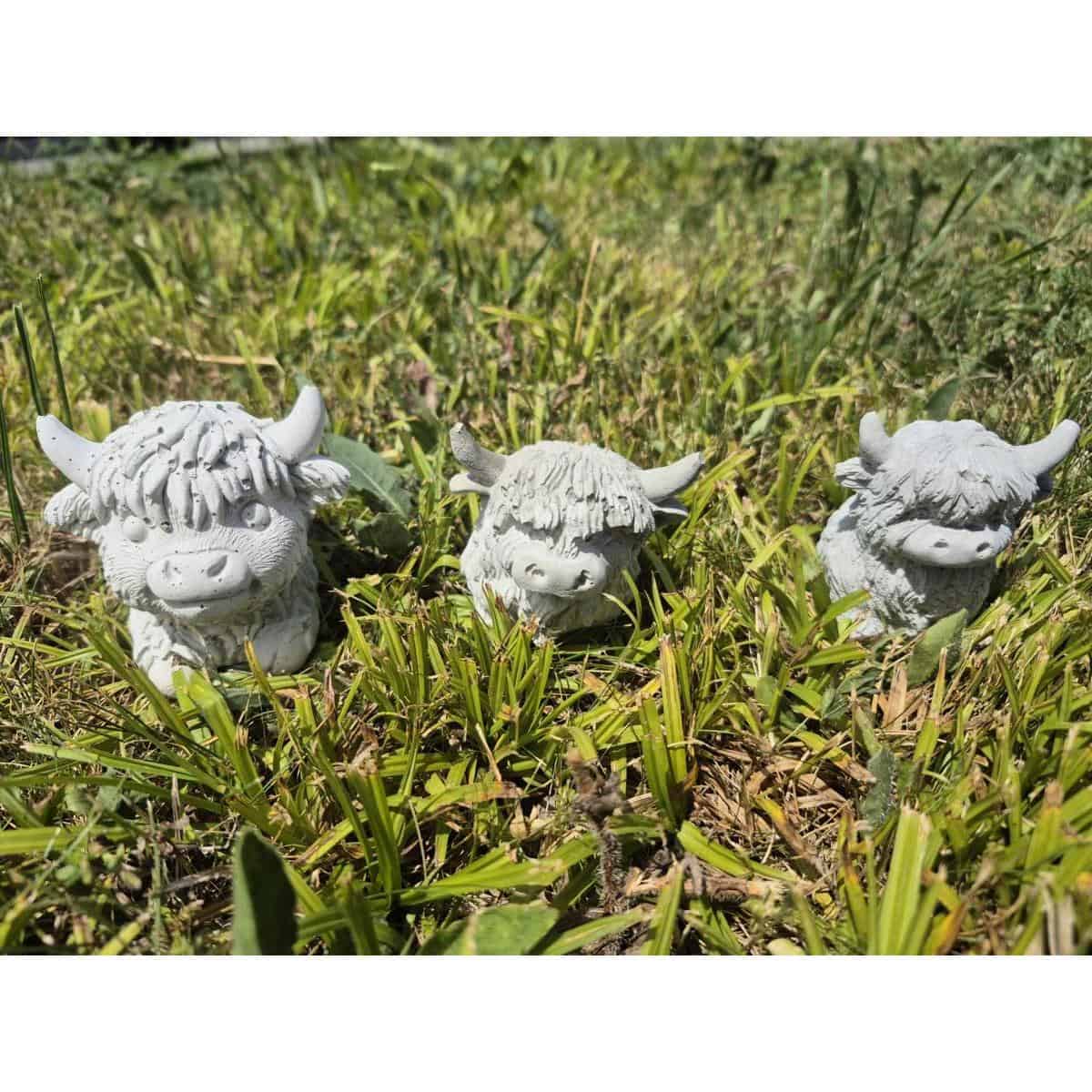 Drei kleine, weiße Spüllappen (Kopie) – Hochlandkuh-Figuren aus Keramik – sitzen im strahlenden Sonnenlicht auf dem Gras.