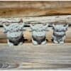 Drei kleine Kuh-Steinstatuen, die Hochlandkühe darstellen, stehen auf einer Holzfläche vor einer rustikalen Holzwand.