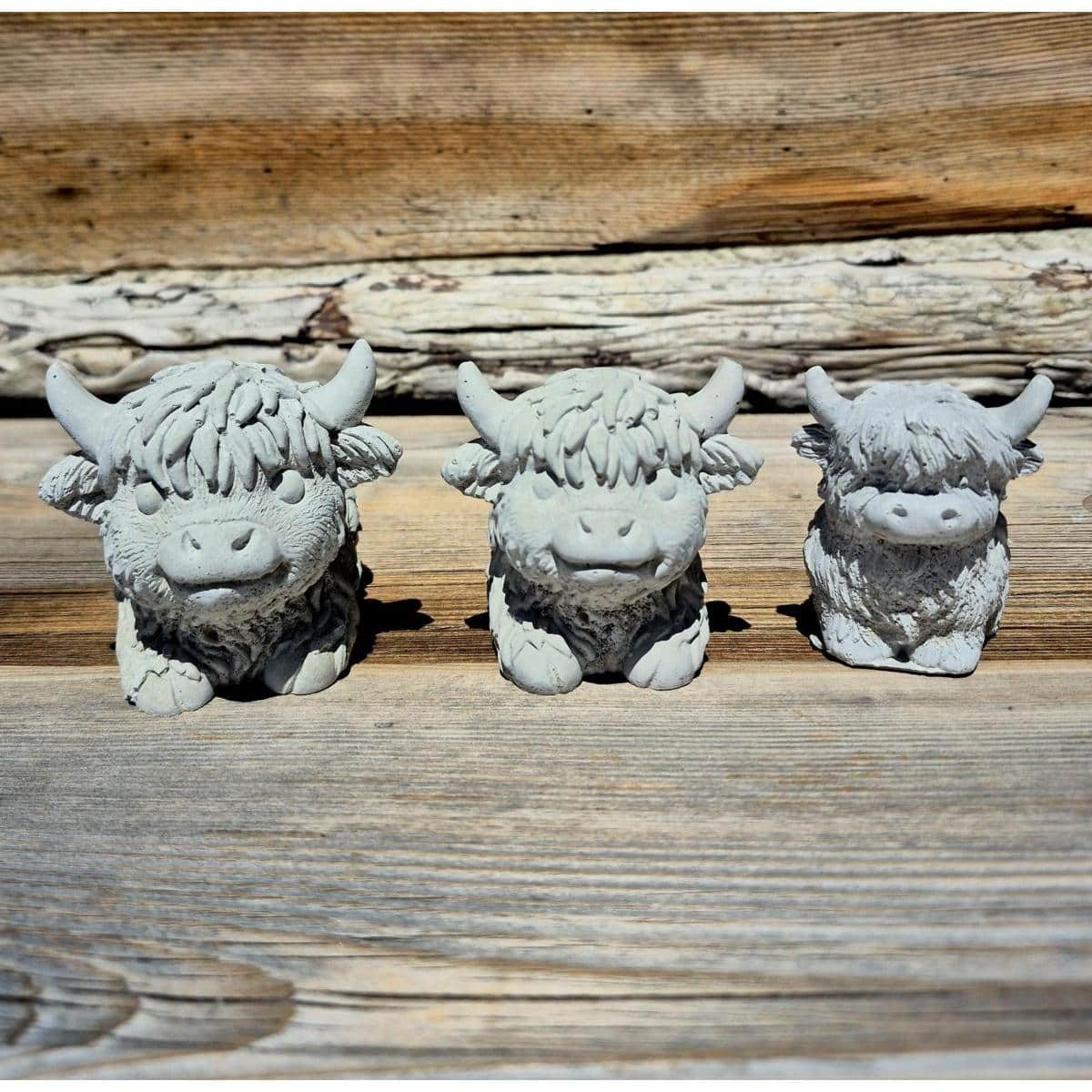 Drei kleine Kuh-Steinstatuen, die Hochlandkühe darstellen, stehen auf einer Holzfläche vor einer rustikalen Holzwand.