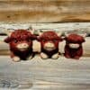 Drei kleine Kuh-Figuren – rote Hochlandkühe aus Ton mit großen Augen –, die auf einer Holzfläche vor einem hölzernen Hintergrund angeordnet sind.